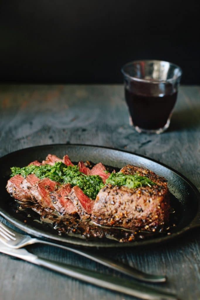 Cast iron skillet with steak chimichurri and a glass of red wine on a rustic table