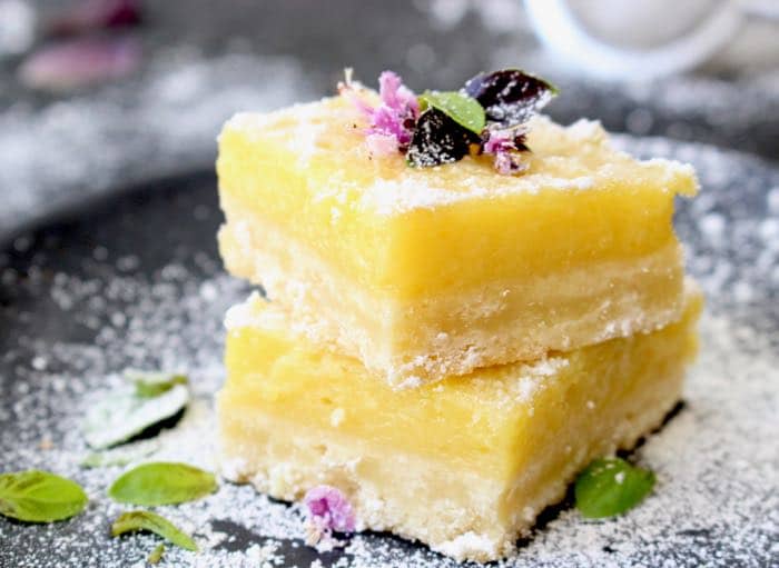 Bright yellow lemon bars with purple basil blossoms on a black rustic table